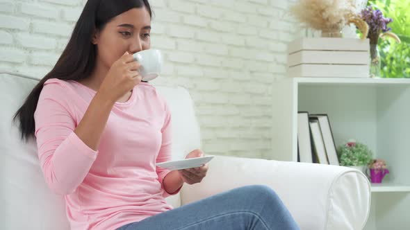 asian young woman drinking warm coffee or tea enjoying it while sitting in her living room at home.