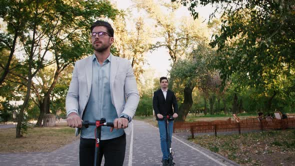 Portraits of Two Handsome Young Businessman Riding an Electric Scooters on the Road in City Park