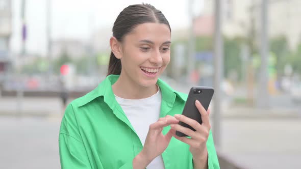 Hispanic Woman Celebrating Online Success on Smartphone Outdoor