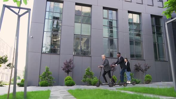 Positive Modern Young Multiracial Friends which Riding on Electric Scooters Near Beautiful Building