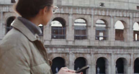 A Woman Dials a Message on Her Mobile Phone While Standing in Front of Coliseum