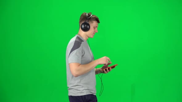 Man in Big Headphones Goes and Dances with Smartphone on Green Screen at Studio. Side View. Slow