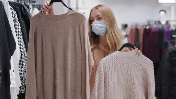 Young Girl in Medical Mask Choose Clothes in Store Woman Consumer Make Choice Between Two Sweaters