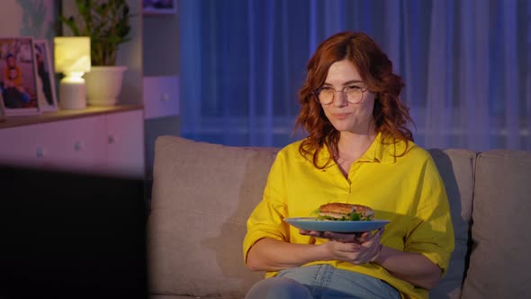 Adult Girl with Glasses Watches Funny Show and Eats Hamburger During an Evening Rest at Home in