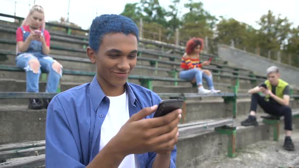 Smiling Male Student Chatting by Smartphone App on Classmates Phones Background
