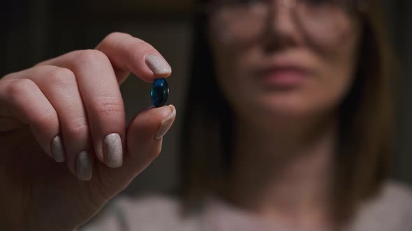 Young Woman Take Blue Capsule and Drinking a Glass of Water Sitting in Dark Room in Low Key