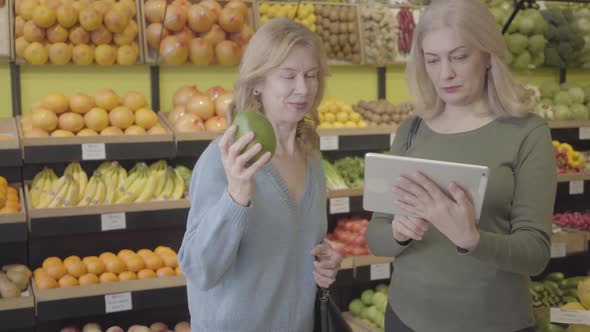 Confident Blond Caucasian Woman with Tablet Advising Female Friend Choice of Best Fruit in Grocery