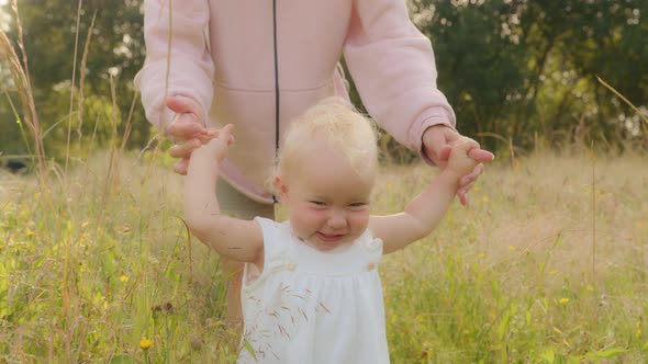 Happy Toddler Smiling Walks Through Meadow in Green High Grass Holding Unrecognizable Mother By Hand