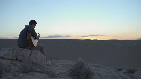 Guy plays the guitaron a hill during desert sunset