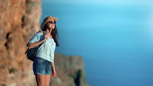 Beautiful Smiling Relaxed Young Woman in Hat and Sunglasses with Backpack Admiring Amazing Sea