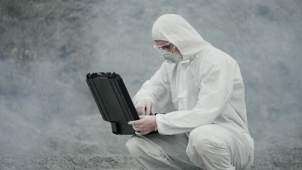 Lab technician in a mask and chemical protective suit opens a box around toxic smoke