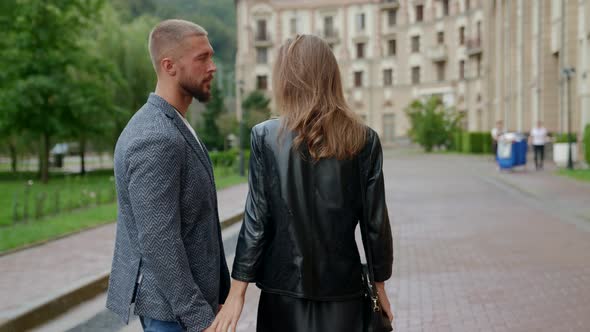 a Man with a Beard and a Blonde in a Black Jacket Walk Along the Path Between the Hotel and the Park