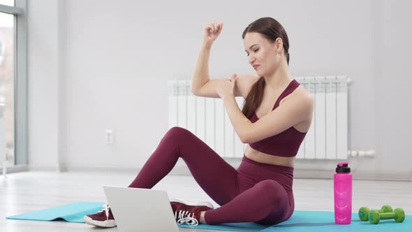 a Young Woman in a Burgundy Tracksuit with Laptop on a Mat in a Gym