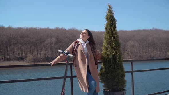 A Young Student Girl in Demiseason Clothes with an Electric Scooter Stands on an Observation Deck By