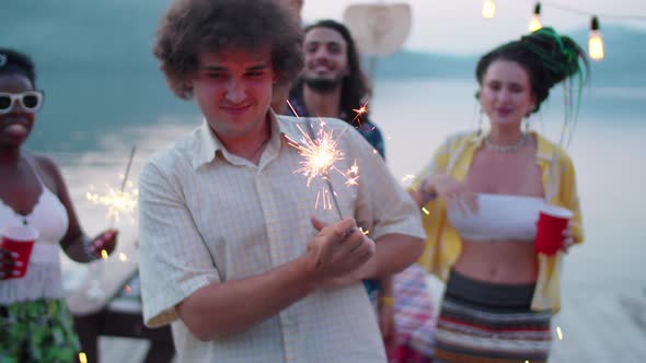 Young Happy Man Dancing with Sparkler at Lake Party with Friends