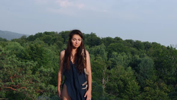 Beautiful Wild Medieval Woman in a Blue Dress Interwoven Feathers in Hair Stands Against of Forest