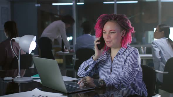 Portrait of Smiling Businesswoman with Pink Hair Talk on Cellphone and Work on Laptop in Dark Office