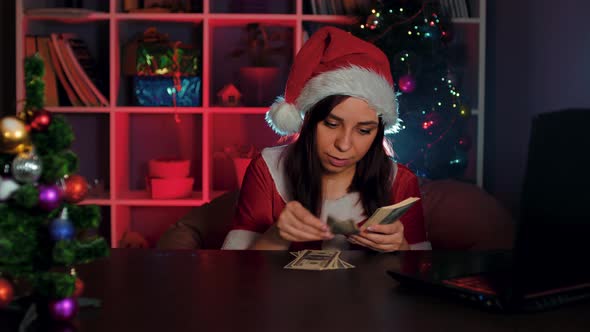 A Young Woman in a Santa Claus Costume Counts Money While Sitting in a Chair in Front of a Computer