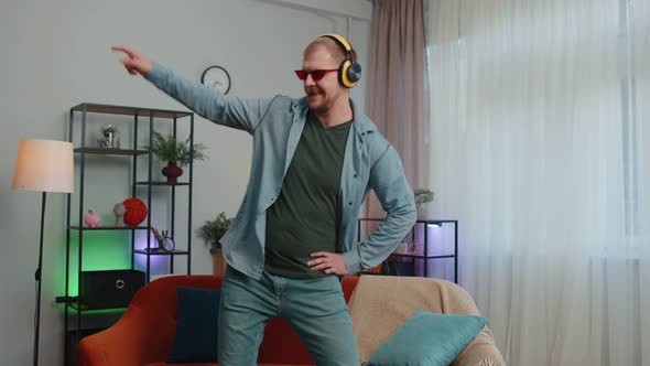 Overjoyed Young Man in Wireless Headphones Dancing Singing on Cozy Couch in Living Room at Home