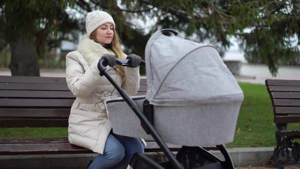 Young Mother with Newborn Child Outdoor. She Sitting on the Bench with Baby Sleeping in Pram