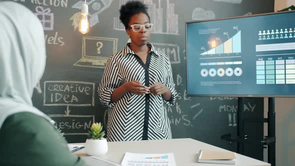 AfroAmerican Businesswoman Talking to Female Muslim Colleague Making Report Pointing at Digital