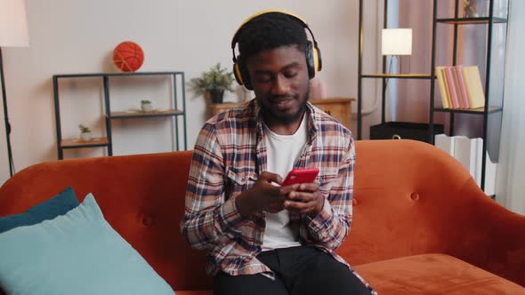 Overjoyed Young Man in Wireless Headphones Dancing Singing on Cozy Couch in Living Room at Home