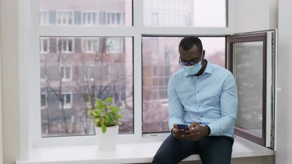 African American Male Business Man in a Medical Mask Holds a Smartphone in His Hands in an Office
