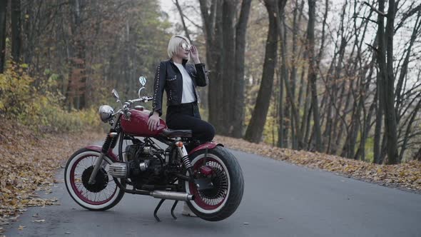 Portrait of Confident Blonde Poses and Corrects Sunglasses at Motorbike