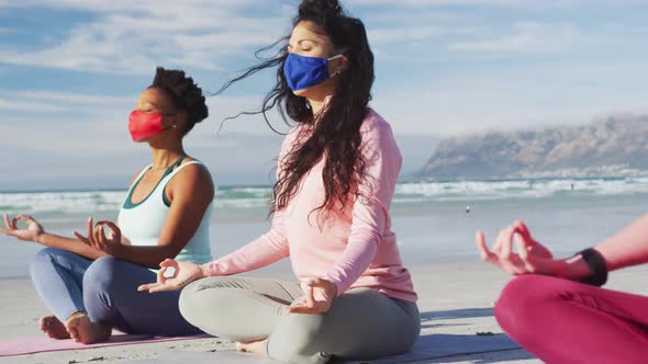 Group of diverse female friends wearing face masks meditating at the beach