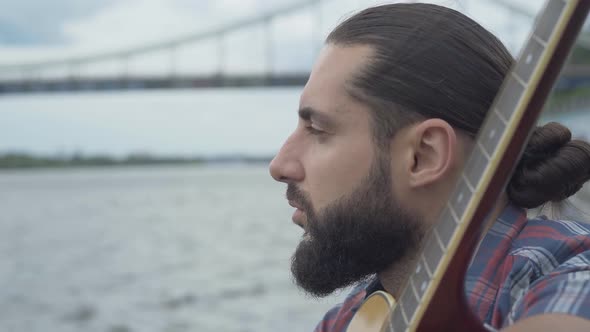 Close-up Face of Thoughtful Artist Sitting with Guitar on River Bank. Side View of Caucasian