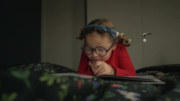 Little Girl in Glasses Lying on the Bed and Reading Book