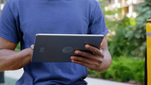 Close Up Afroamerican Man Courier Food Delivery Sitting on Bench Uses Tablet