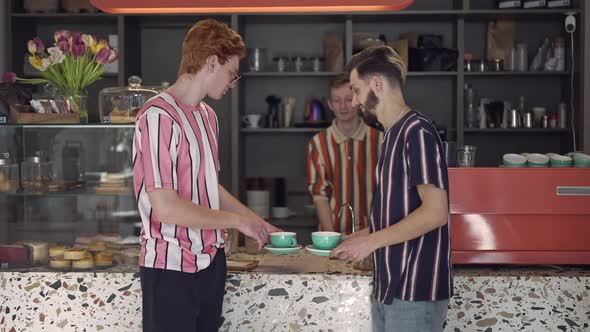 Male Barista Pushing Coffee Cups to Clients in Vintage Coffee Shop As Satisfied Young Caucasian Men