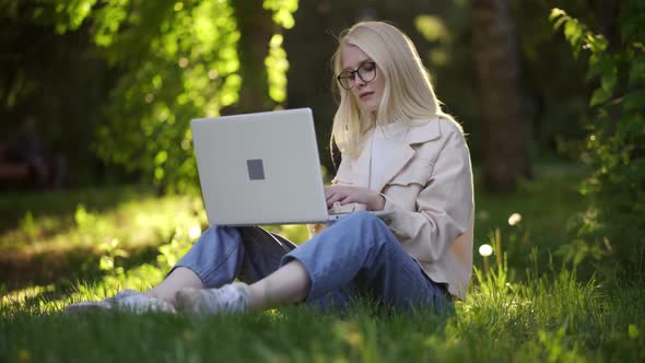 A Young Blonde Woman with a Laptop Sits on a Green Meadow in the Park