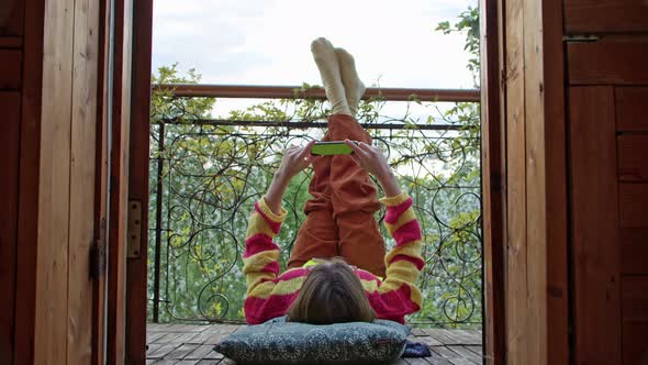 Carefree Woman Lies on the Balcony and Looks Into Her Smartphone Moving Her Legs for Joy