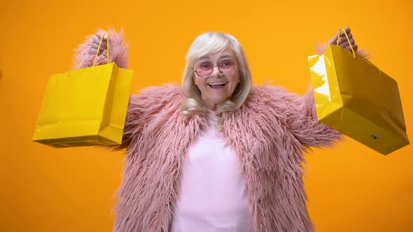 Happy Aging Lady in Funny Pink Coat Showing Yellow Shopping Bags, Wasting Money