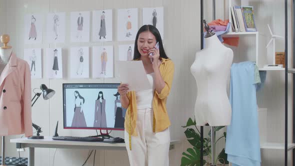 Designer Talking On Smartphone, Looking At Picture In Hand And Walking Around While Design Clothes