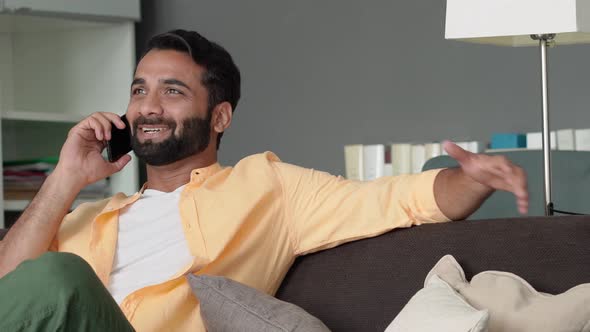 Happy Indian Man Sitting at Home on Sofa Talking on Cell Phone