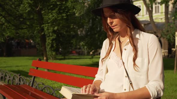 Young Beautiful Girl in Hat Reading Book in City Park