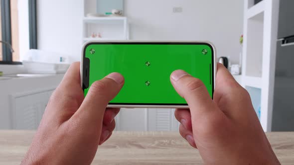 Man's Hand Playing App Game on Smartphone With Green Mockup Screen Chroma Key on Kitchen Background