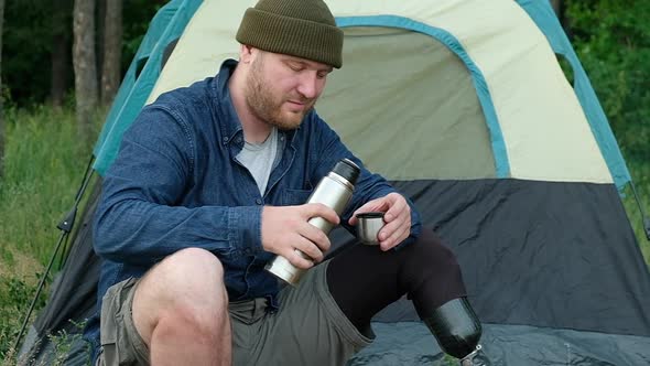 disabled person in  forest is sitting by fire and drinking tea from thermos