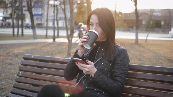 Beautiful Woman Use Phone Sitting on a Park Bench with Coffee Cup.