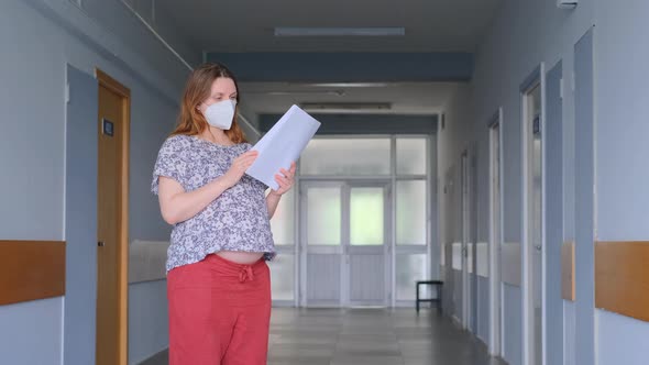 Pregnant woman stands with papers and documents in the clinic with a face mask