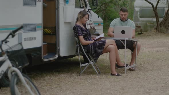 Man working at laptop and woman reading the book in summer camping.