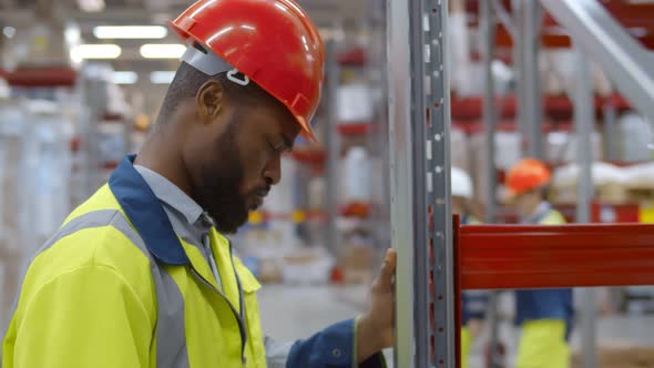 Exhausted African Worker Hitting Head in Hardhat Against Shelf in Warehouse