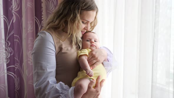Happy Young Mother Holds Baby Girl in Arms Kissing By Window