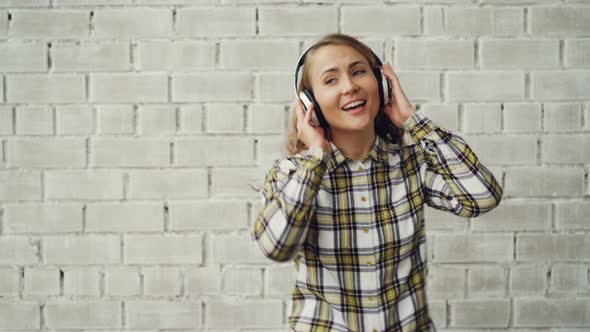 Portrait of Attractive Girl Listening To Music Through Headphones, Singing and Dancing Against Brick