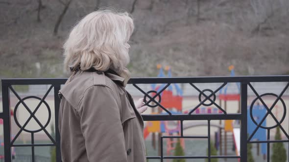 Side View of Thoughtful Senior Caucasian Woman Standing Next To Fence and Looking Away. Blond Mature