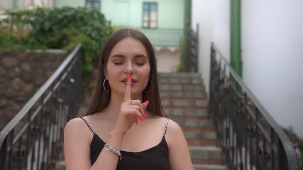 Young pretty girl on the background of the old town bringing index finger to her lips.