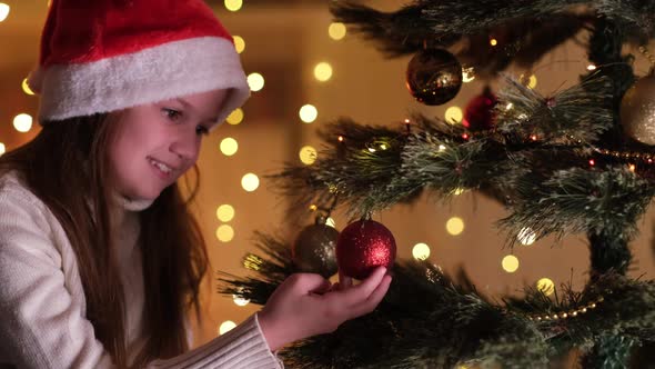 Cute Little Girl in White Sweater and Santa Hat Holding Decorative Toy Ball on Christmas Tree Branch